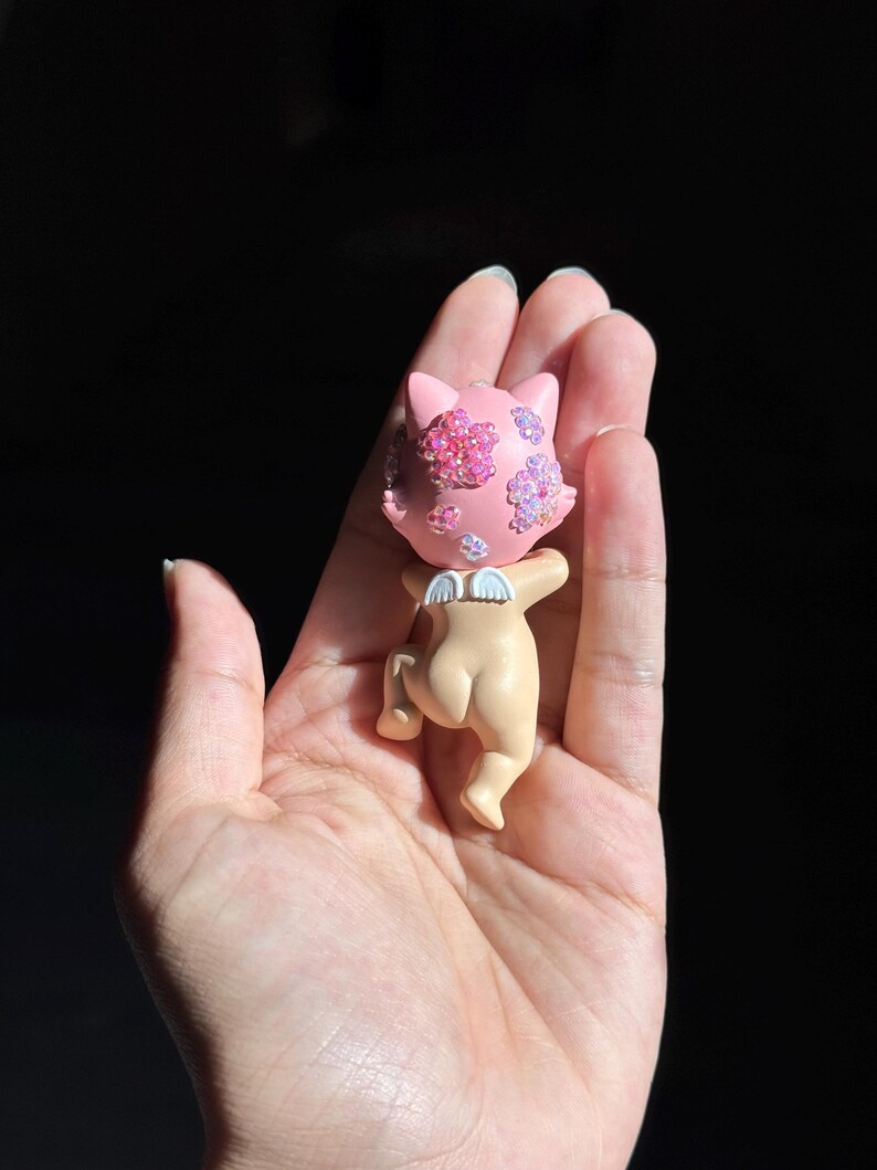 May include: A small figurine of a cat-like creature with a light pink head adorned with iridescent floral-shaped crystals. The figure has a tan-colored body, small white wings on its back, and is depicted in a crawling position with its head turned.