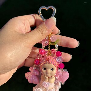 May include: A pink fairy keychain with a gold heart-shaped clasp. The fairy figurine has a sweet expression and is adorned with pink beads, flowers, and a delicate dress. The keychain is a charming accessory.