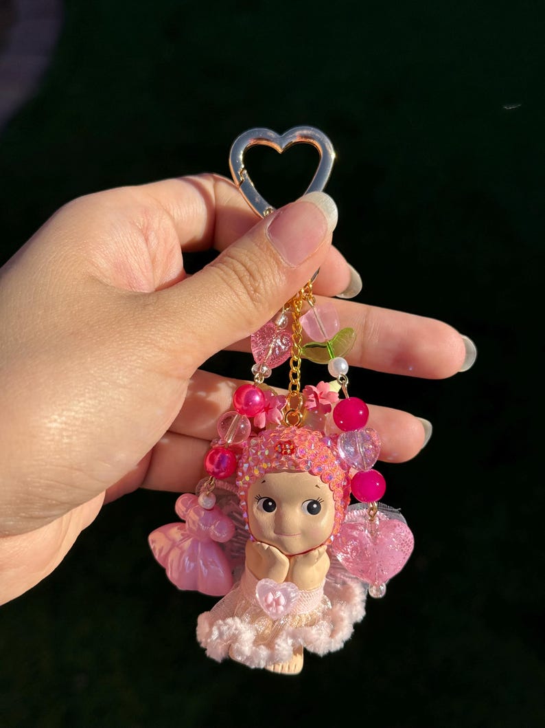 May include: A pink fairy keychain with a gold heart-shaped clasp. The fairy figurine has a sweet expression and is adorned with pink beads, flowers, and a delicate dress. The keychain is a charming accessory.