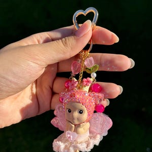 May include: A pink fairy keychain with a gold heart-shaped clasp. The fairy figurine has a pink jeweled crown, wings, and a ruffled skirt. The keychain also features pink heart and flower charms.