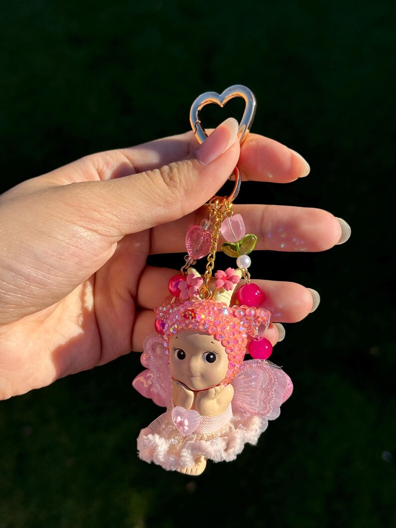 May include: A pink fairy keychain with a gold heart-shaped clasp. The fairy figurine has a pink jeweled crown, wings, and a ruffled skirt. The keychain also features pink heart and flower charms.