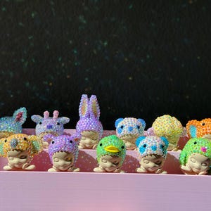 May include: A collection of eleven small figurines, each featuring a face with closed eyes and wearing a different animal-themed hood covered in sparkling, multi-colored rhinestones, arranged in two rows on a light pink tiered display stand.