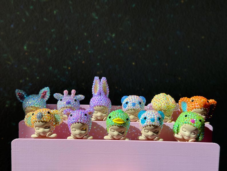 May include: A collection of eleven small figurines, each featuring a face with closed eyes and wearing a different animal-themed hood covered in sparkling, multi-colored rhinestones, arranged in two rows on a light pink tiered display stand.