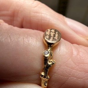 Peut inclure: Une bague dor&eacute;e avec une bande textur&eacute;e et un petit dessus rond. Le dessus de la bague pr&eacute;sente un motif grav&eacute;. Une petite pierre pr&eacute;cieuse transparente est sertie dans la bande. La bague est sur un doigt.