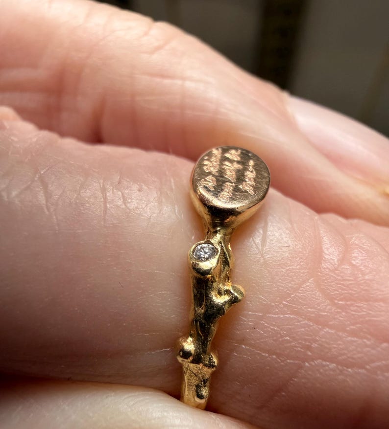 Peut inclure: Une bague dor&eacute;e avec une bande textur&eacute;e et un petit dessus rond. Le dessus de la bague pr&eacute;sente un motif grav&eacute;. Une petite pierre pr&eacute;cieuse transparente est sertie dans la bande. La bague est sur un doigt.