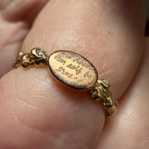 Peut inclure: Une bague en or avec une plaque ovale grav&eacute;e des mots "To thine own self be true". La bague a un anneau textur&eacute; avec de petits d&eacute;tails ronds et deux petits diamants.