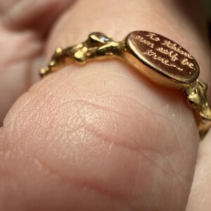 Peut inclure: Une bague en or avec une pi&egrave;ce ma&icirc;tresse de forme ovale. La bague a un anneau textur&eacute; avec de petites bosses irr&eacute;guli&egrave;res. L'ovale porte l'inscription "To thine own self be true..." en cursive. Une petite pierre transparente est sertie pr&egrave;s de l'ovale.