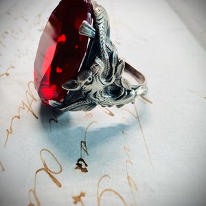 May include: A silver ring featuring a large, faceted, oval-shaped deep red gemstone. The metal band is intricately sculpted with detailed relief carvings of fish or aquatic creatures that wrap around the sides of the central red stone setting.