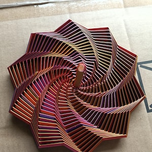 May include: A star-shaped sculpture with a spiral design, featuring layers of metal. The colour gradient includes red, orange, and purple hues. A wooden dowel is at the centre. The sculpture is a decorative object.