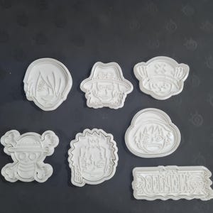 以下が含まれることがあります： アニメ「ワンピース」のキャラクターとシンボルが描かれた白いプラスチック製のクッキーカッターのコレクション。セットには、さまざまなキャラクターの顔、ドクロ、シリーズのロゴが含まれています。