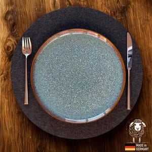 Könnte beinhalten: Ein tealfarbener, gesprenkelter Keramikteller mit kupferfarbenem Rand liegt auf einem dunkelgrauen Filz-Tischset. Eine silberne Gabel und ein Messer sind auf beiden Seiten des Tellers platziert. Der Teller steht auf einer Holzoberfläche. Mit "Made in Germany" Logo.