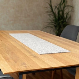 Könnte beinhalten: Ein rechteckiger Esstisch aus hellem Holz mit einem grauen Steineinsatz. Der Tisch wird von einem Holzrahmen mit schwarzen Metallakzenten getragen. Vier graue, gepolsterte Stühle umgeben den Tisch. Der Tisch ist etwa 180 cm lang.