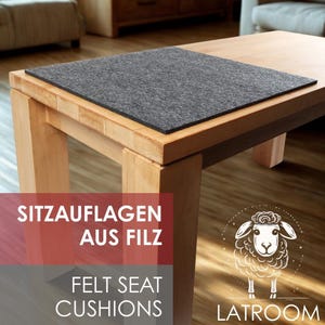 Pode incluir: Uma almofada de assento retangular de feltro cinza escuro repousa sobre uma mesa de madeira clara. A mesa tem um design simples e moderno. A imagem também inclui o texto "FELT SEAT CUSHIONS" e um logotipo de ovelha.