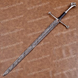 May include: A long, ornate sword with a patterned blade and a silver hilt. The handle is wrapped in black material. The sword features a crossguard and a decorative pommel. The background is a brown leather-like surface.