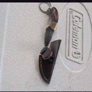 May include: A small fixed-blade knife with a dark wood handle, a brass bolster, and a patterned steel blade. The knife is attached to a metal keyring and is accompanied by a dark brown leather sheath featuring a snap closure and visible stitching.