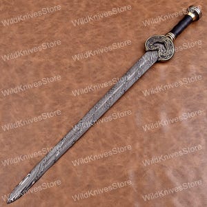 May include: A decorative sword with a patterned blade and ornate hilt. The hilt features a black grip and gold-colored accents. The blade has a unique, swirling pattern. The background is a brown, textured surface. The text "WildKnivesStore" is visible.
