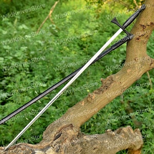 May include: A long, black and silver sword with a cross-shaped hilt and a pointed blade. The sword is resting on a tree branch against a backdrop of green foliage. The handle has a textured grip.