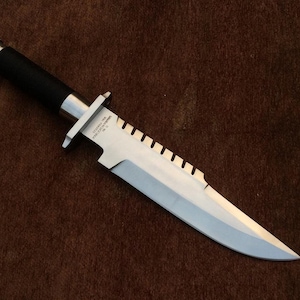 May include: A large, stainless steel knife with a black handle and a serrated edge. The knife has a silver guard and a pointed blade. The text on the blade reads "STAINLESS USA MARBLES GLADSTONE, MI. NO. 12".