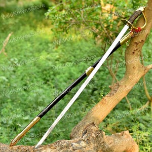 May include: A saber with a curved, polished steel blade and a black handle. The hilt is gold-colored with a curved guard. The scabbard is black with gold accents. The sword is displayed on a tree branch against a green background.