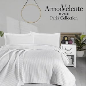 Op de afbeelding: Witte beddengoedset met een gestructureerd, gestreept patroon. De set bevat een dekbedovertrek en kussenslopen. De afbeelding toont ook een nachtkastje met decoratieve items en een plant. De tekst "ArmonVelente HOME Paris Collection" is zichtbaar.