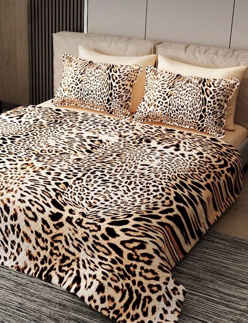 Puede incluir: Juego de cama con estampado de leopardo. El conjunto incluye una manta y dos fundas de almohada. El dise&ntilde;o presenta manchas negras sobre un fondo beige. La ropa de cama est&aacute; colocada sobre una cama con cabecero de color neutro.