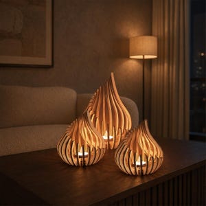 Puede incluir: Tres portavelas de madera de color claro con un diseño de láminas en forma de lágrima. Cada portavelas presenta láminas de madera verticales dispuestas en una estructura curva y abierta, con una pequeña vela encendida en el centro de cada pieza.