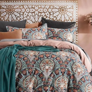 Op de afbeelding: Een bed met een patroon dekbedovertrek, kussens en een turquoise sprei. Het dekbedovertrek en de kussenslopen hebben een bloemenpatroon in tinten van turquoise, oranje en crème. Een gesneden houten hoofdbord met een geometrisch patroon is ook zichtbaar.