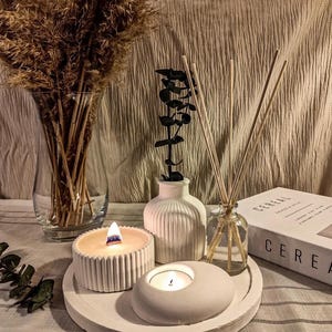 Op de afbeelding: Een rustgevende opstelling met een brandende kaars met een houten lont, een kleine kaars en een rietverspreider. De opstelling omvat een vaas met gedroogde planten, een witte vaas met een plant en een boek. Het kleurenpalet is neutraal met zachte verlichting.