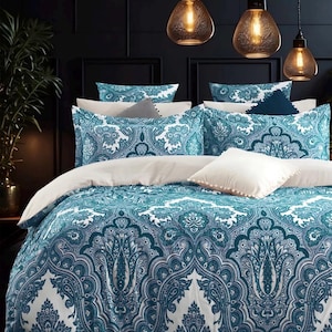Pode incluir: Conjunto de roupa de cama com um padrão paisley turquesa e branco. O conjunto inclui uma capa de edredão, fronhas e almofadas decorativas. O design é detalhado e ornamentado. O fundo apresenta uma parede escura e lâmpadas suspensas.