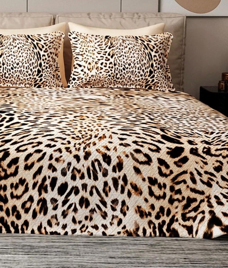 Puede incluir: Juego de cama con estampado de leopardo. El conjunto incluye una funda n&oacute;rdica y dos fundas de almohada. El dise&ntilde;o presenta manchas marrones y negras sobre un fondo beige, creando una est&eacute;tica salvaje y ex&oacute;tica. La ropa de cama est&aacute; colocada sobre una cama.