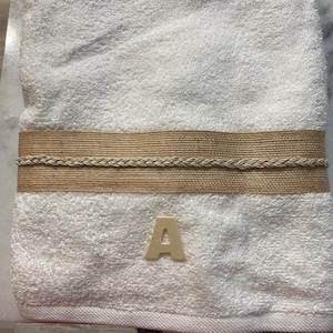 May include: White bath towel with a decorative band of tan fabric and braided trim. A wooden letter "A" is placed on the towel. The towel is folded and appears soft and plush.
