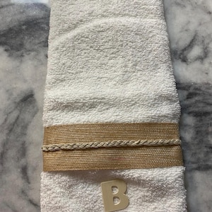 May include: White hand towel with a decorative band of tan rope and a braided rope accent. A small, beige letter "B" is affixed to the bottom of the towel. The towel is made of a soft, absorbent material.