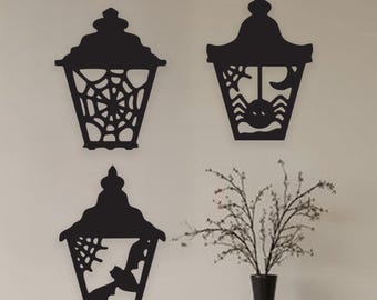 May include: Three black Halloween lantern wall decorations. Each lantern features a different cutout design: a spider web, a spider with a crescent moon, and a bat with a spider web. A black vase with branches sits on a shelf.