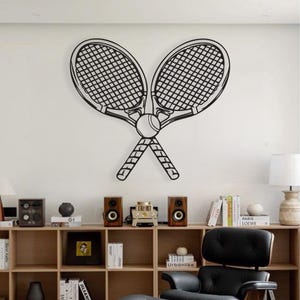 Puede incluir: Arte de pared de metal negro con dos raquetas de tenis cruzadas y una pelota de tenis. Las raquetas tienen un patrón de rejilla, y el diseño está montado en una pared blanca. Ideal para los amantes del deporte.