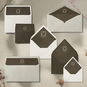 Op de afbeelding: Een assortiment enveloppen in verschillende maten en vormen. De enveloppen zijn wit en donkerbruin, met een decoratief wapen gedrukt op de flap en binnenkant. Ze zijn gerangschikt op een neutrale achtergrond.