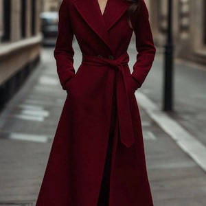 May include: A long, burgundy coat with a belted waist and a classic lapel collar. The coat features long sleeves and pockets, creating a sophisticated and elegant silhouette. The coat is made of a soft, flowing fabric.