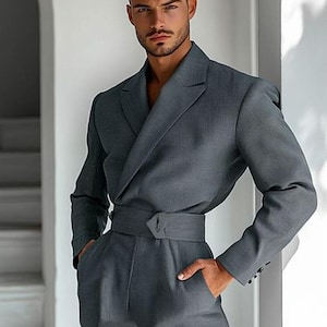 May include: A gray suit with a wrap-around belt. The suit has a double-breasted jacket and matching trousers. The model is leaning against a white wall with his hands in his pockets.