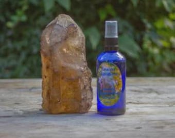 May include: A large, rough-cut, amber-colored crystal stands next to a cobalt blue spray bottle. The bottle has a silver cap and a colorful label. The background is a blurred view of green foliage.