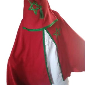 May include: A red hooded cloak with green trim and embroidered star details. The cloak has a wide, flowing design and is paired with a white garment underneath. The hood features a green tassel.