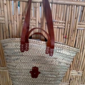 May include: Woven straw tote bag with brown leather handles and a small brown leather Hamsa hand emblem. The bag has a rectangular shape and a woven pattern. The handles are attached with decorative stitching.