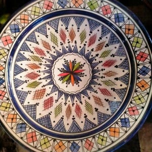 May include: A decorative ceramic plate with a vibrant, hand-painted design. The plate features a central starburst pattern in multiple colors, surrounded by diamond and square motifs in blue, orange, green, and red. A dark blue border frames the design.