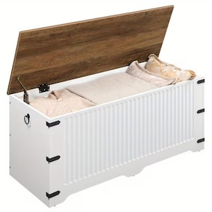 May include: A white rectangular storage chest with a hinged wooden lid featuring a natural wood grain finish. The exterior has vertical grooved paneling, black metal corner accents, and side ring handles. The chest is filled with folded beige textiles.