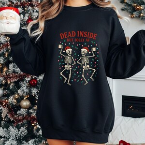 May include: Black sweatshirt with the text "DEAD INSIDE BUT JOLLY AF" above a graphic of two skeletons wearing Santa hats and holding mugs. The skeletons are surrounded by Christmas lights.