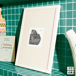 Könnte beinhalten: Ein gerahmter Druck mit einem Schwarz-Weiß-Linolschnitt eines Reptilienkopfes. Der Druck befindet sich in einem weißen Rahmen und lehnt an einer grünen Fliesenoberfläche. Ein Buch mit dem Titel "Fucking Fernand" ist sichtbar.
