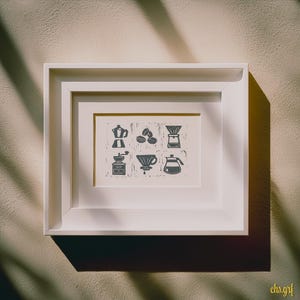 Könnte beinhalten: Ein gerahmter Druck mit Kaffee-Illustrationen in einem weißen Rahmen. Der Druck zeigt eine Kaffeemaschine, Kaffeebohnen, einen Pour-Over, eine Mühle, einen Dripper und eine Kaffeekanne. Das Kunstwerk ist in Grau- und Weißtönen gehalten, mit der Signatur des Künstlers unten rechts.
