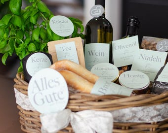 May include: A wicker gift basket filled with various gourmet food items. The basket contains a bottle of wine, olive oil, pasta, honey, breadsticks, and tea. Green herbs and decorative labels add to the presentation. A white ribbon adorns the front.