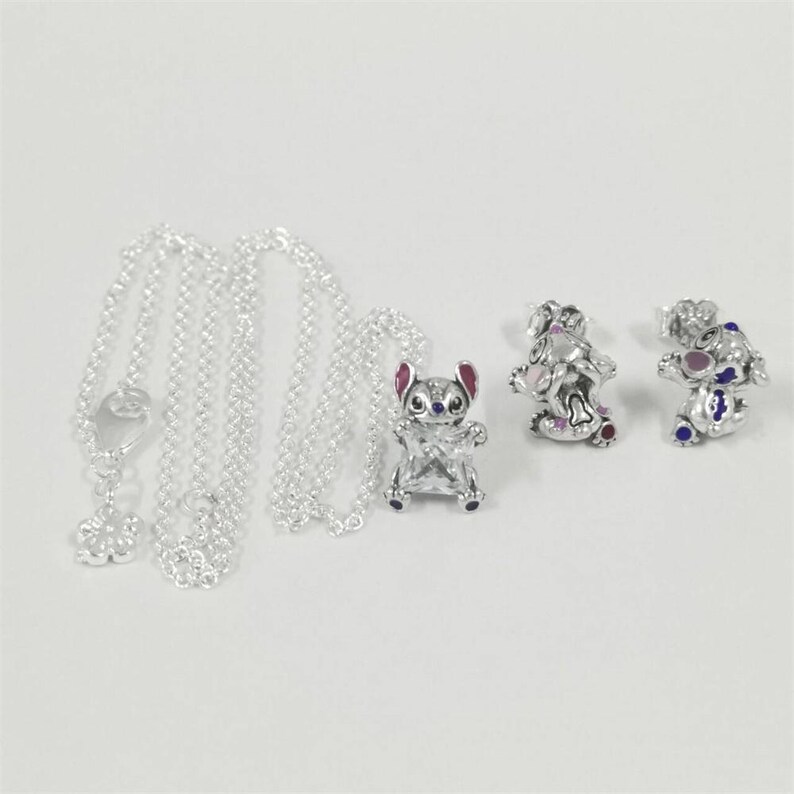 May include: A set of silver-coloured jewellery including a necklace with a flower charm, a pendant, and a pair of earrings. The pendant and earrings are shaped like a cartoon character with pink ears and blue accents.