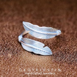 May include: A silver ring with a feather design. The ring features two detailed feather accents. The ring is set against a brown background. The text "OKOYEJUSTIN Handcrafted Jewellery" is at the bottom. The ring is a piece of handcrafted jewellery.
