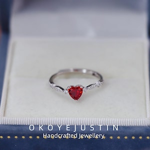 May include: A silver ring with a heart-shaped, red gemstone. The ring features a delicate, twisted band with small, clear accent stones. The ring is displayed in a blue jewelry box. The text "OKOYEJUSTIN Handcrafted Jewellery" is visible.