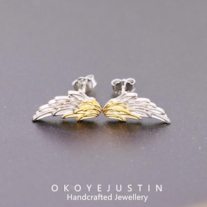 Puede incluir: Un par de pendientes de botón plateados y dorados con forma de ala. Los pendientes tienen un diseño detallado de plumas con una capa exterior plateada y un centro dorado. La marca "OKOYEJUSTIN Handcrafted Jewellery" está en la parte inferior.